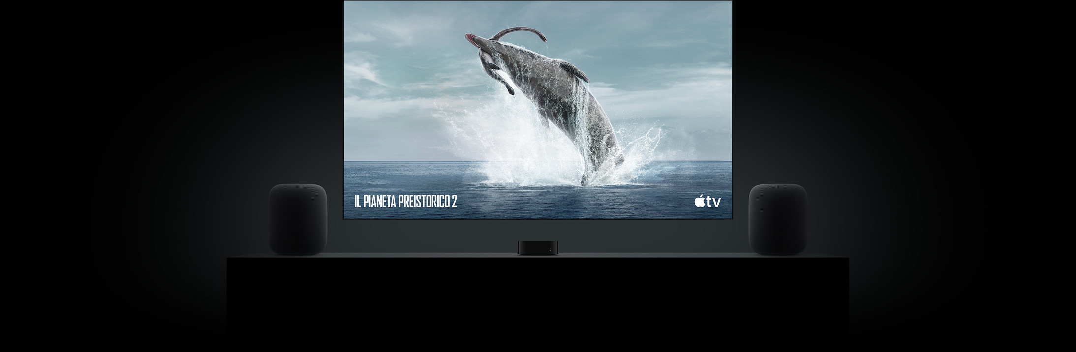 Un grande televisore a schermo piatto con l’immagine brillante di un dinosauro tratta da Il pianeta preistorico. Il televisore è appeso al muro sopra a un mobile da salotto con una Apple TV al centro e due HomePod ai lati