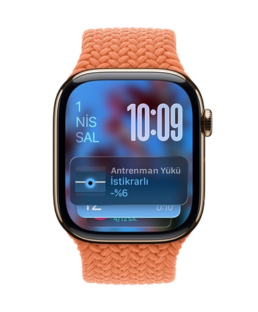 Koyu Turuncu örgü kayışa sahip Roze Altın rengi Apple Watch'un kadranında sol üstte tarih, sağ üstte sıralanmış rakamlarla saat, altta Antrenman Yükü araç takımı yer alıyor; hepsi yarı saydam Liquid Glass görünümünde