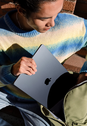 นักศึกษานั่งอยู่นอกอาคาร พร้อมหยิบ MacBook ออกจากกระเป๋าเป้