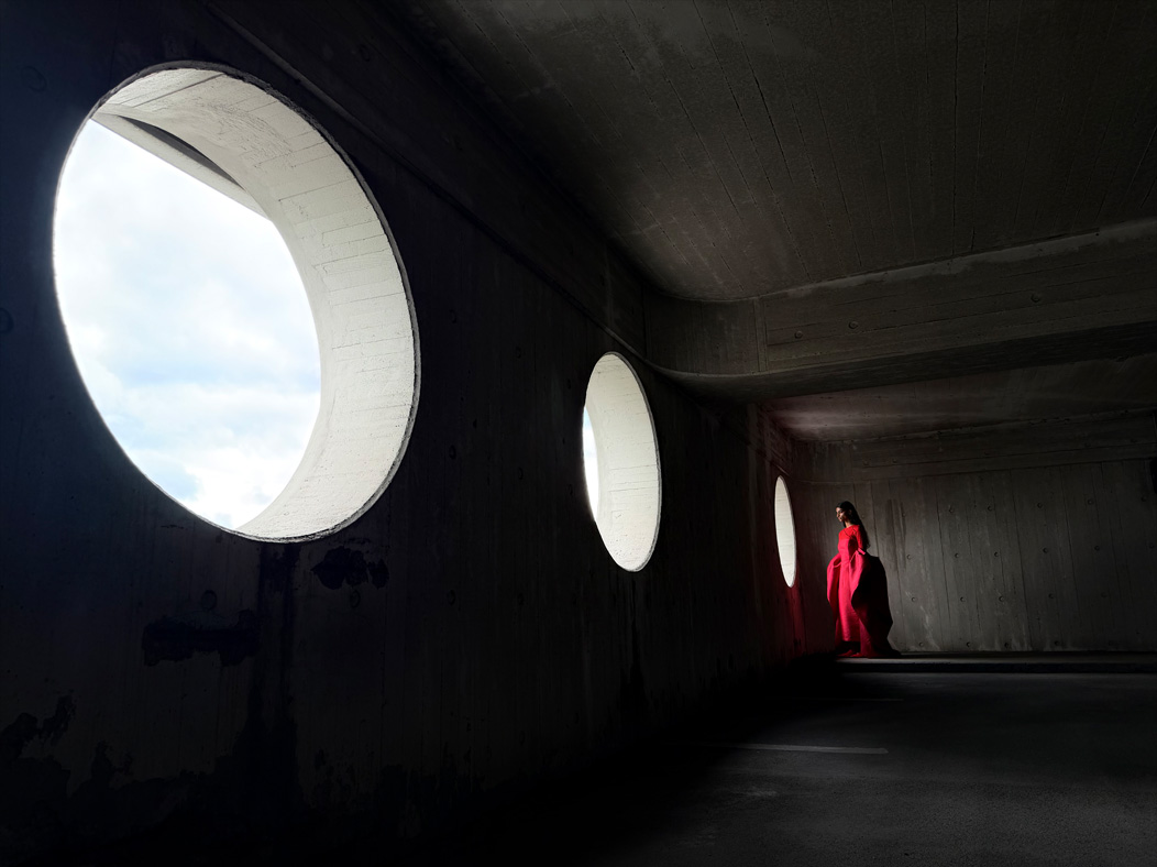 Uma mulher com um longo vestido vermelho a ser fotografada numa sala vazia. A app Fotografia muda de distância focal para ilustrar as capacidades do sistema de câmaras Pro, a mostrar a opção de 24 mm
