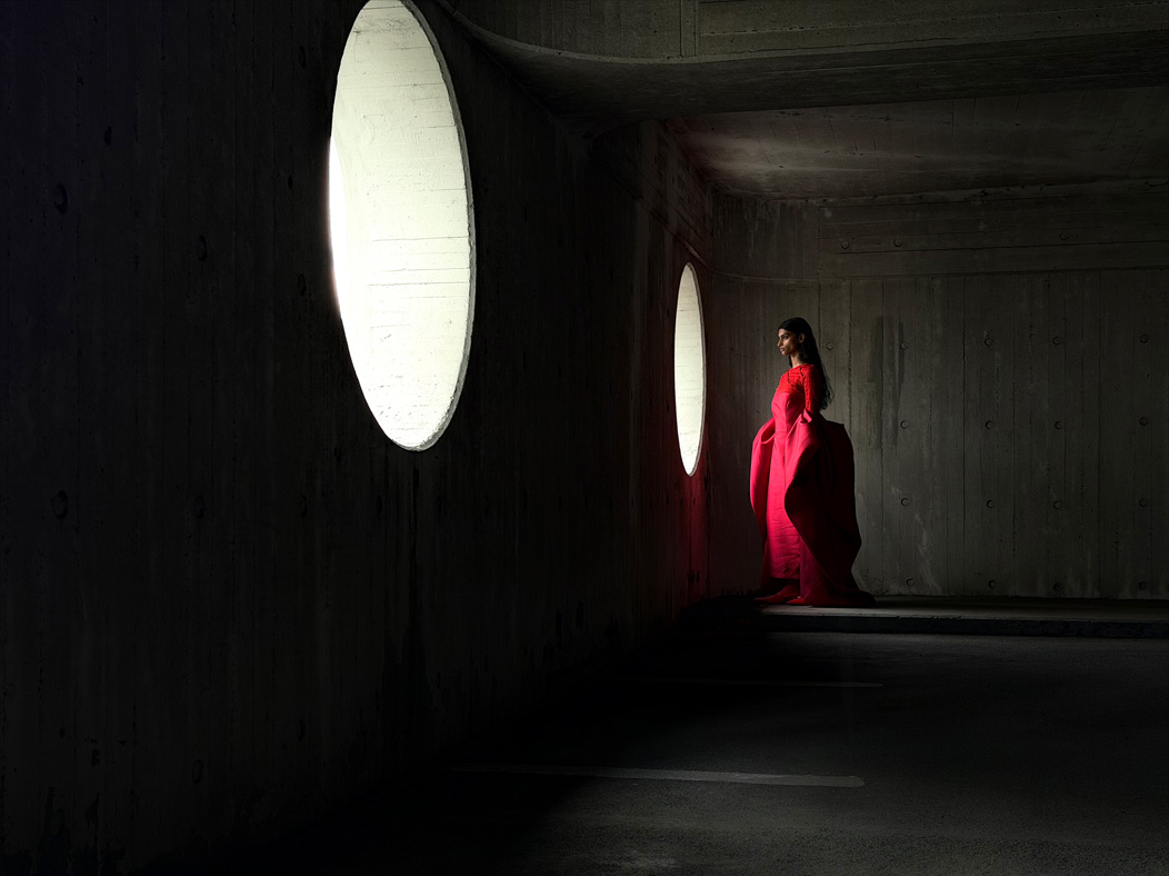 Uma mulher com um longo vestido vermelho a ser fotografada numa sala vazia. A app Fotografia muda de distância focal para ilustrar as capacidades do sistema de câmaras Pro, a mostrar a opção de 48 mm